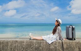 Eine junge Frau in Sommerkleidung und mit Sonnenhut sitzt auf einer kleinen Mauer neben dem Meer, neben ihr steht ein Koffer.