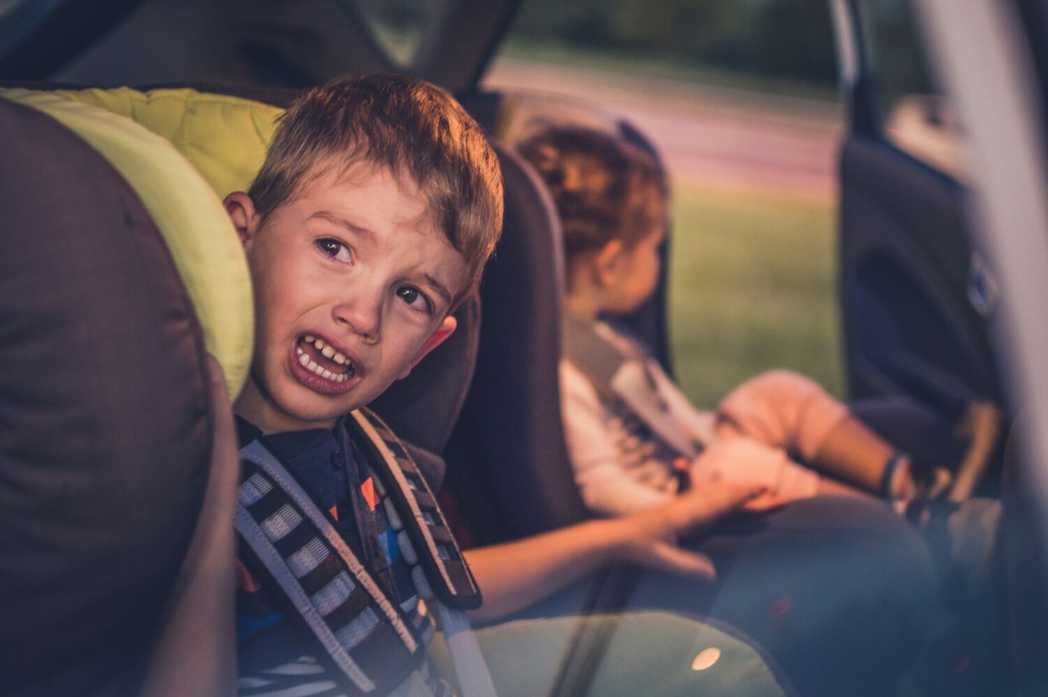 Zwei in ihren Kindersitzen gesicherte Kinder in einem Auto. Das ältere Kind hat einen quengelnden Gesichtsausdruck.