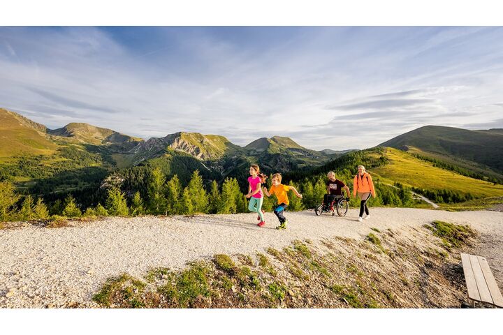Wanderweg in den Bergen in Kärnten. Zwei Kinder laufen vor, die Erwachsenen, ein Mann im Rollstuhl und eine Frau folgen ihnen.