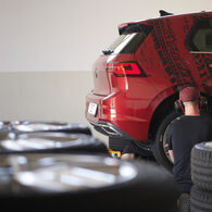 ÖAMTC-Studie: Reifenabrieb 2025 Auf dem Bild ist der hintere Teil eines roten Autos zu sehen. Ein Mann mit roter Mütze und Kopfhörern kniet mit dem Rücken zur Kamera vor dem Hinterrad des Autos. Im Vordergrund, zum Teil verschwommen: Mehrere Stapel Autoreifen.