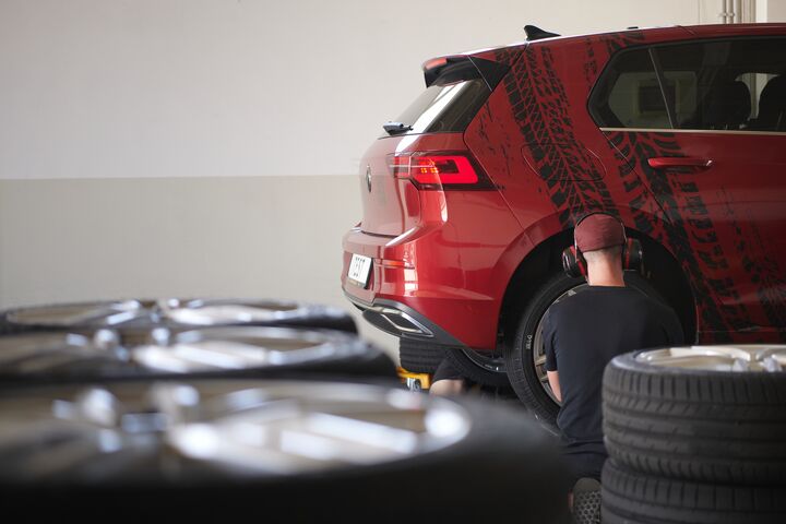 Auf dem Bild ist der hintere Teil eines roten Autos zu sehen. Ein Mann mit roter Mütze und Kopfhörern kniet mit dem Rücken zur Kamera vor dem Hinterrad des Autos. Im Vordergrund, zum Teil verschwommen: Mehrere Stapel Autoreifen.