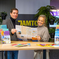 Das Bild zeigt zwei Personen, die hinter einem Tisch stehen auf dem viele Reise-Broschüren liegen. In den Händen halten die zwei Personen eine aufgeklappte Reisekarte.