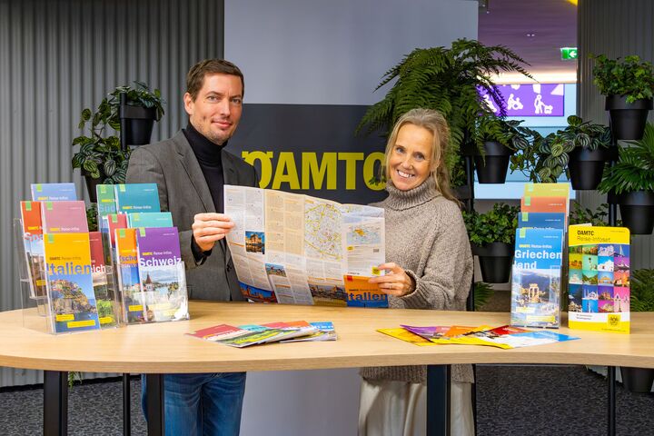 Das Bild zeigt zwei Personen, die hinter einem Tisch stehen auf dem viele Reise-Broschüren liegen. In den Händen halten die zwei Personen eine aufgeklappte Reisekarte.