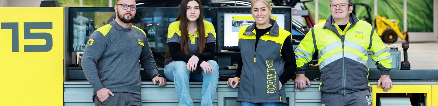 Vier ÖAMTC-Mitarbeitende stehen und sitzen in einer Werkstatt vor einer Prüfstraße, im Hintergrund ist ein Auto auf einer Hebebühne zu sehen.