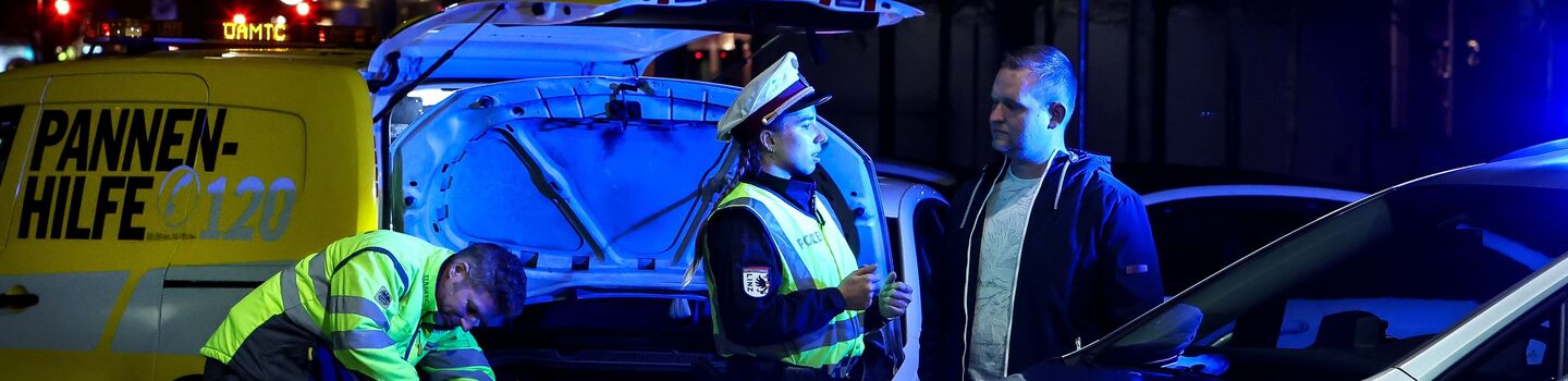 Polizistin klärt Autofahrenden über die Wichtigkeit einer ausreichenden Beleuchtung auf. Im Hintergrund wird von einem ÖAMTC-Techniker die Lampe beim Auto getauscht. Die gemeinsame Aktion nennt sich Licht statt Strafe.