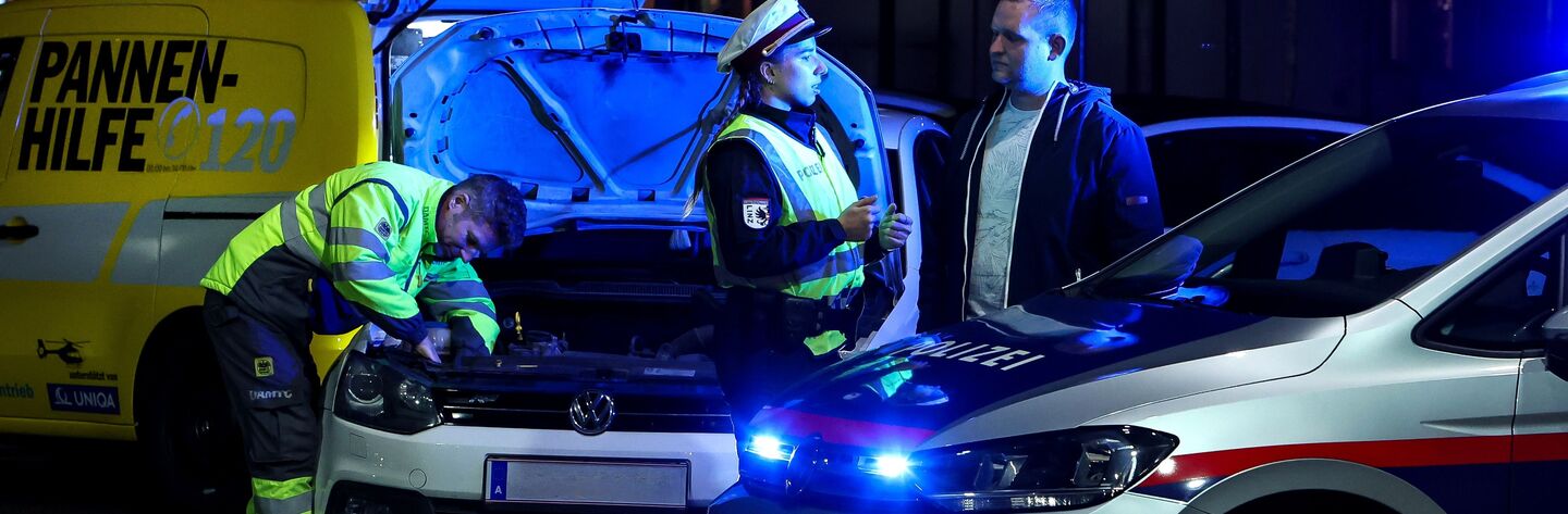 Polizistin klärt Autofahrenden über die Wichtigkeit einer ausreichenden Beleuchtung auf. Im Hintergrund wird von einem ÖAMTC-Techniker die Lampe beim Auto getauscht. Die gemeinsame Aktion nennt sich Licht statt Strafe.