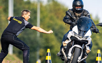 0219_24 Header_FT_Website_1920x900.jpg Ein Motorradfahrer fährt auf einem Übungsgelände. Der Fahrer trägt eine komplette Schutzausrüstung, einschließlich Helm, Jacke und Handschuhe. Links und rechts neben dem Fahrer stehen Verkehrshütchen. Links neben dem Motorradfahrer steht ein Trainer und gibt Anweisungen.