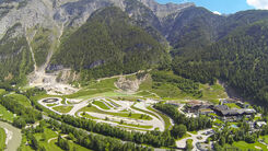 Inmitten einer grünen und bergigen Landschaft erstreckt sich das Fahrtechnikzentrum Saalfelden-Brandlhof. Eine große asphaltierte Übungsfläche, mit Kreuzungselementen, Bewässerungsanlagen, Bürogebäuden und einem Hotel. Im Hintergrund heben sich die Alpen mit bewaldeten Berghängen und felsigen Gipfeln.