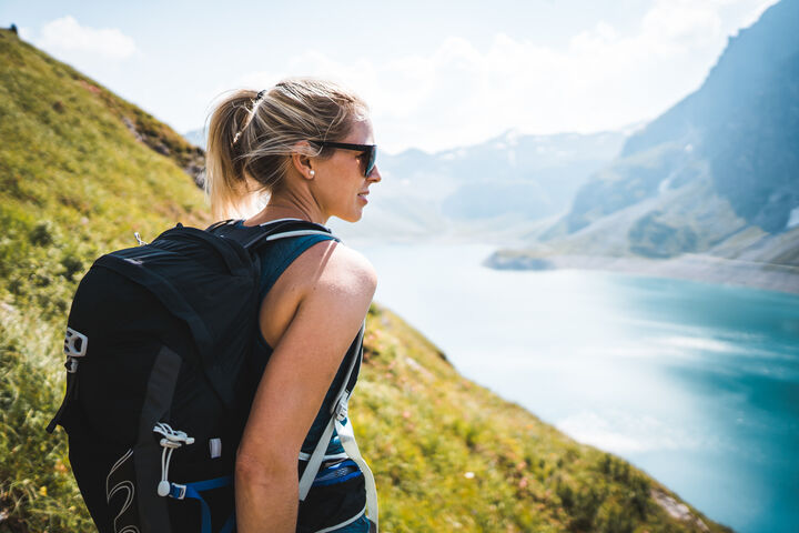 Eine Frau mit Sonnenbrille und Rucksack steht vor einem See in einer bergigen Landschaft.