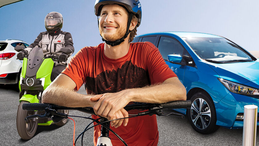 Ein Radfahrer mit Helm, ein E-Mopedfahrer mit Helm und ein blaues E-Auto beim Laden an einer Tankstelle. Im Hintergrund ist eine Wasserstoff Lademöglichkeit.