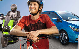 Ein Radfahrer mit Helm, ein E-Mopedfahrer mit Helm und ein blaues E-Auto beim Laden an einer Tankstelle. Im Hintergrund ist eine Wasserstoff Lademöglichkeit.