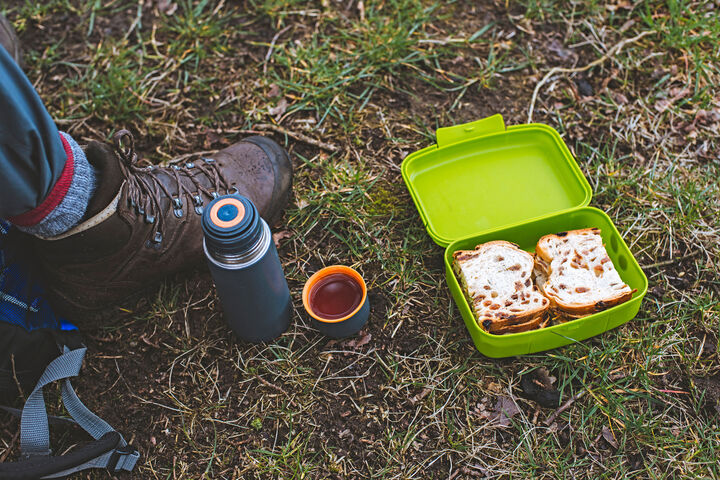 Eine Jausenbox befüllt mit einem Brot und eine Trinlflasche liegen in einer Wiese neben dem Fuß einer Person.