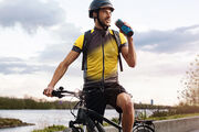 Ein Radfahrer in gelb-schwarzer Fahrradkleidung und Helm sitzt auf einem Fahrrad und trinkt aus einer Wasserflasche. Er befindet sich auf einem Radweg neben einem Fluss.
