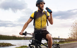 Ein Radfahrer in gelb-schwarzer Fahrradkleidung und Helm sitzt auf einem Fahrrad und trinkt aus einer Wasserflasche. Er befindet sich auf einem Radweg neben einem Fluss.