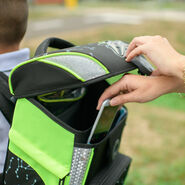 Ein Junge trägt einen Schultasche die offen ist. Man sieht eine Hand, die ein Handy in die Schultasche steckt.