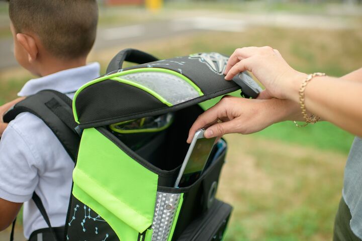 Ein Junge trägt einen Schultasche die offen ist. Man sieht eine Hand, die ein Handy in die Schultasche steckt.