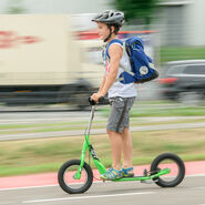 Ein Junge fährt auf einem grünen Tretroller, auch Scooter genannt, auf einem Weg neben einer Straße. Er trägt einen Helm und eine blaue Schultasche am Rücken.