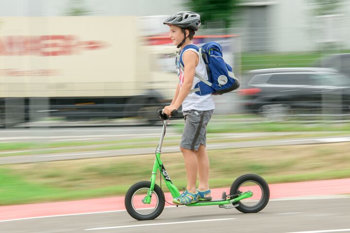 Ein Junge fährt auf einem grünen Tretroller, auch Scooter genannt, auf einem Weg neben einer Straße. Er trägt einen Helm und eine blaue Schultasche am Rücken.