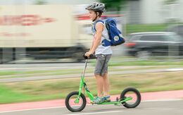 Ein Junge fährt auf einem grünen Tretroller, auch Scooter genannt, auf einem Weg neben einer Straße. Er trägt einen Helm und eine blaue Schultasche am Rücken.