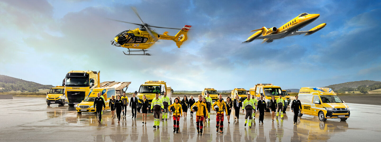 Eine Flotte von gelben Rettungsfahrzeugen und Luftfahrzeugen steht auf einem Flugfeld. Zu sehen ein Krankenwagen, ÖAMTC Pannenfahrzeuge, ein Abschlepp-LKW, ein Rettungshubschrauber in der Luft sowie ein Ambulanzflugzeug. Davor stehen Einsatzkräfte in leuchtender Schutzkleidung. 