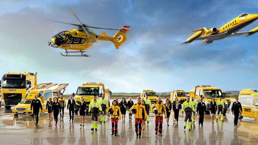 Eine Flotte von gelben Rettungsfahrzeugen und Luftfahrzeugen steht auf einem Flugfeld. Zu sehen ein Krankenwagen, ÖAMTC Pannenfahrzeuge, ein Abschlepp-LKW, ein Rettungshubschrauber in der Luft sowie ein Ambulanzflugzeug. Davor stehen Einsatzkräfte in leuchtender Schutzkleidung.