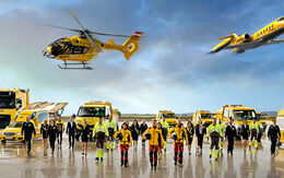 Eine Flotte von gelben Rettungsfahrzeugen und Luftfahrzeugen steht auf einem Flugfeld. Zu sehen ein Krankenwagen, ÖAMTC Pannenfahrzeuge, ein Abschlepp-LKW, ein Rettungshubschrauber in der Luft sowie ein Ambulanzflugzeug. Davor stehen Einsatzkräfte in leuchtender Schutzkleidung.