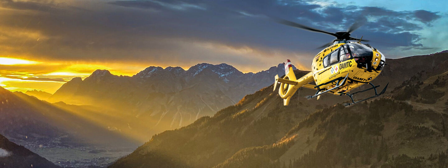 Gelber Hubschrauber der ÖAMTC-Flugrettung fliegt bei Sonnenaufgang im Gebirge.