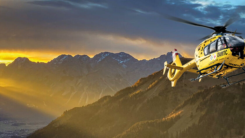 Gelber Hubschrauber der ÖAMTC-Flugrettung fliegt bei Sonnenaufgang im Gebirge.