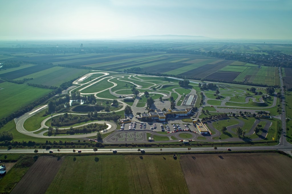 Inmitten einer ländlichen Landschaft erstreckt sich das Fahrtechnik Zentrum Teesdorf. Es gibt viele asphaltierte Trainingsstrecken mit Kreuzungselementen und Kurvenkombinationen, einen Offroad Hügel mit einem Teich, einen Handlingparcours im Motorrad Zentrum, ein Gebäude und einen großen Parkplatz.  Im Hintergrund ist ein Dorf inmitten grüner Felder.  