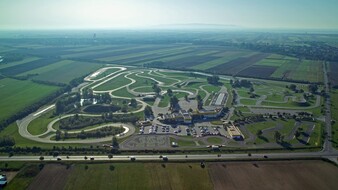 Inmitten einer ländlichen Landschaft erstreckt sich das Fahrtechnik Zentrum Teesdorf. Es gibt viele asphaltierte Trainingsstrecken mit Kreuzungselementen und Kurvenkombinationen, einen Offroad Hügel mit einem Teich, einen Handlingparcours im Motorrad Zentrum, ein Gebäude und einen großen Parkplatz. Im Hintergrund ist ein Dorf inmitten grüner Felder.