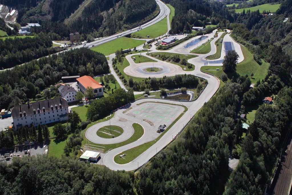 Inmitten einer bewaldeten Landschaft erstreckt sich das Fahrtechnik Zentrum Innsbruck. Es gibt viele asphaltierte Trainingsstrecken mit Kreuzungselementen, Kreisbahn und Kurvenkombinationen. In der Mitte befindet sich ein Teich. Links vom Fahrtechnik Zentrum befinden sich Gebäude.