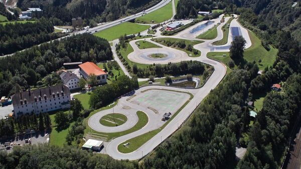 Inmitten einer bewaldeten Landschaft erstreckt sich das Fahrtechnik Zentrum Innsbruck. Es gibt viele asphaltierte Trainingsstrecken mit Kreuzungselementen, Kreisbahn und Kurvenkombinationen. In der Mitte befindet sich ein Teich. Links vom Fahrtechnik Zentrum befinden sich Gebäude.