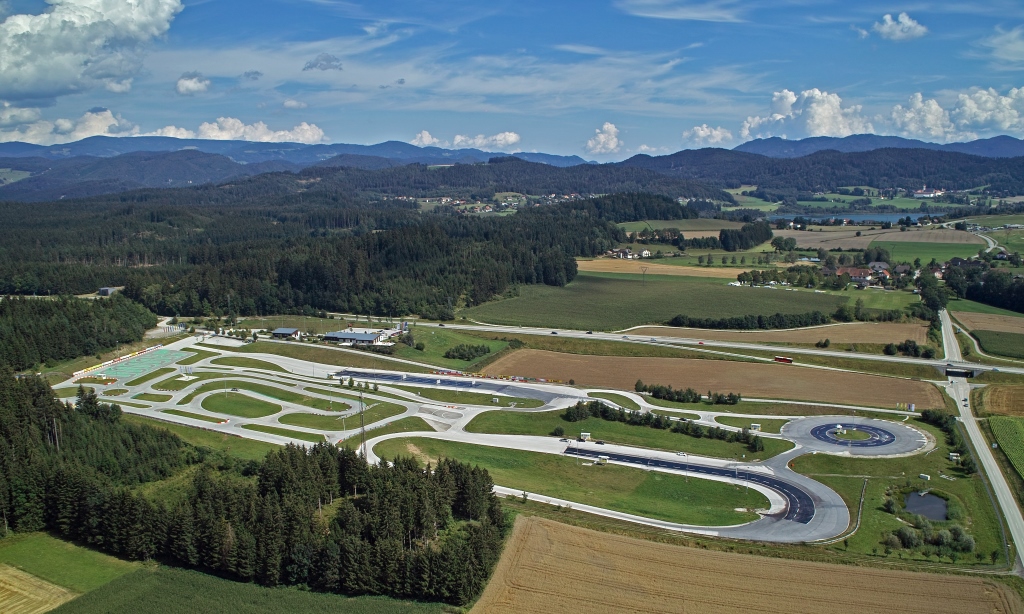 Inmitten einer hügeligen Landschaft erstreckt sich das Fahrtechnik Zentrum St.Veit. Es gibt asphaltierte Trainingsstrecken mit Kreuzungselementen, eine Kreisbahn und Kurvenkombinationen. Im Hintergrund sind Wälder, Häuser und ein See.