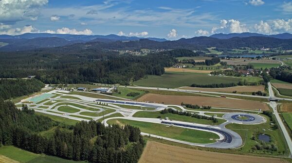 Inmitten einer hügeligen Landschaft erstreckt sich das Fahrtechnik Zentrum St.Veit. Es gibt asphaltierte Trainingsstrecken mit Kreuzungselementen, eine Kreisbahn und Kurvenkombinationen. Im Hintergrund sind Wälder, Häuser und ein See.