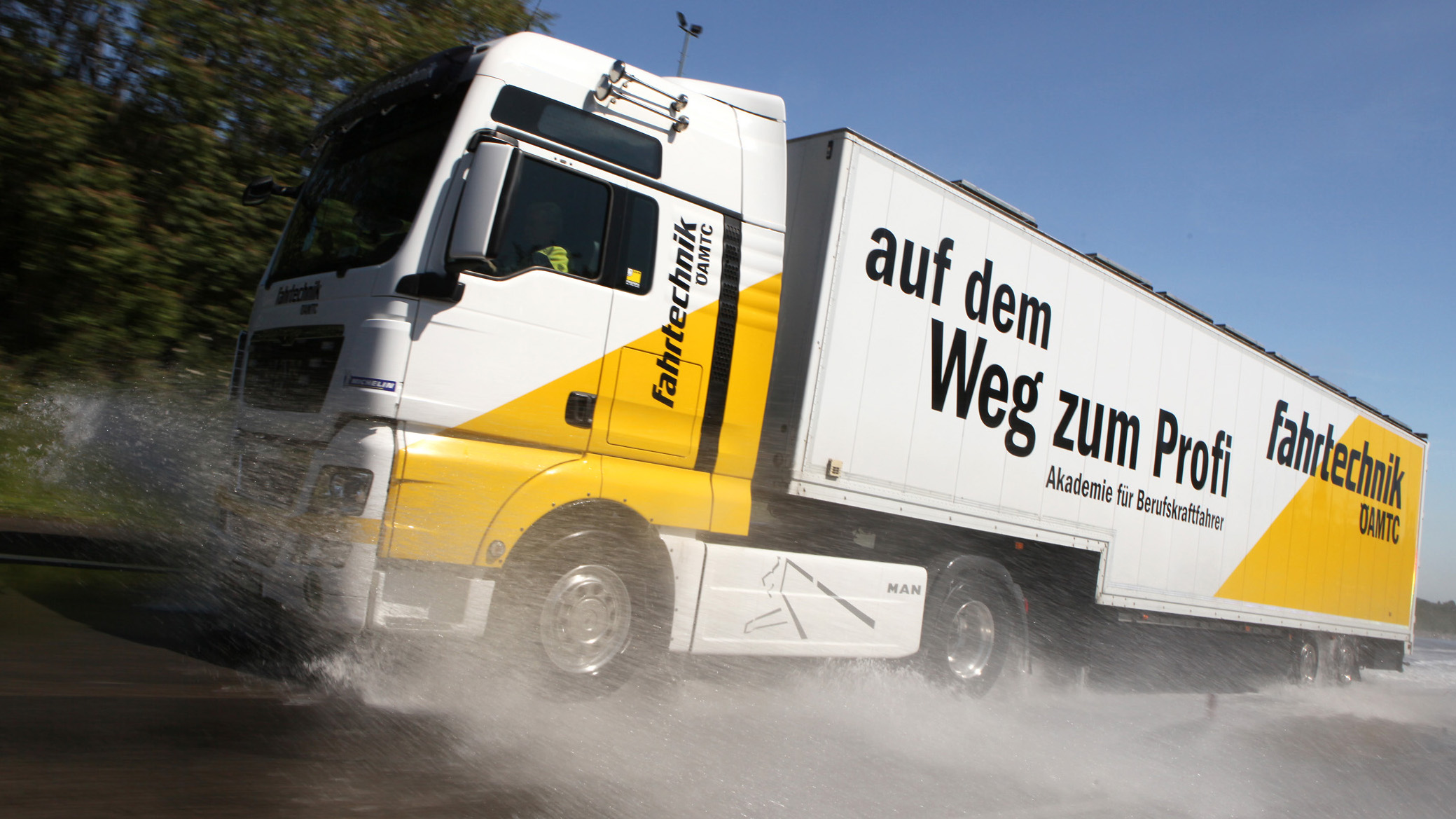 Ein weiß-gelber LKW mit ÖAMTC Fahrtechnik-Beschriftung fährt nach links. Wasser spritzt davon. Im Hintergrund blauer Himmel und Bäume.