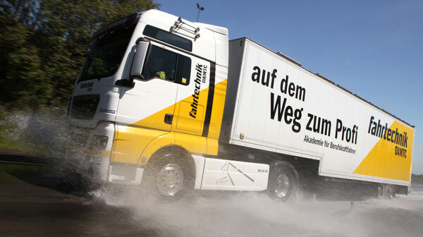 Ein weiß-gelber LKW mit ÖAMTC Fahrtechnik-Beschriftung fährt nach links. Wasser spritzt davon. Im Hintergrund blauer Himmel und Bäume.