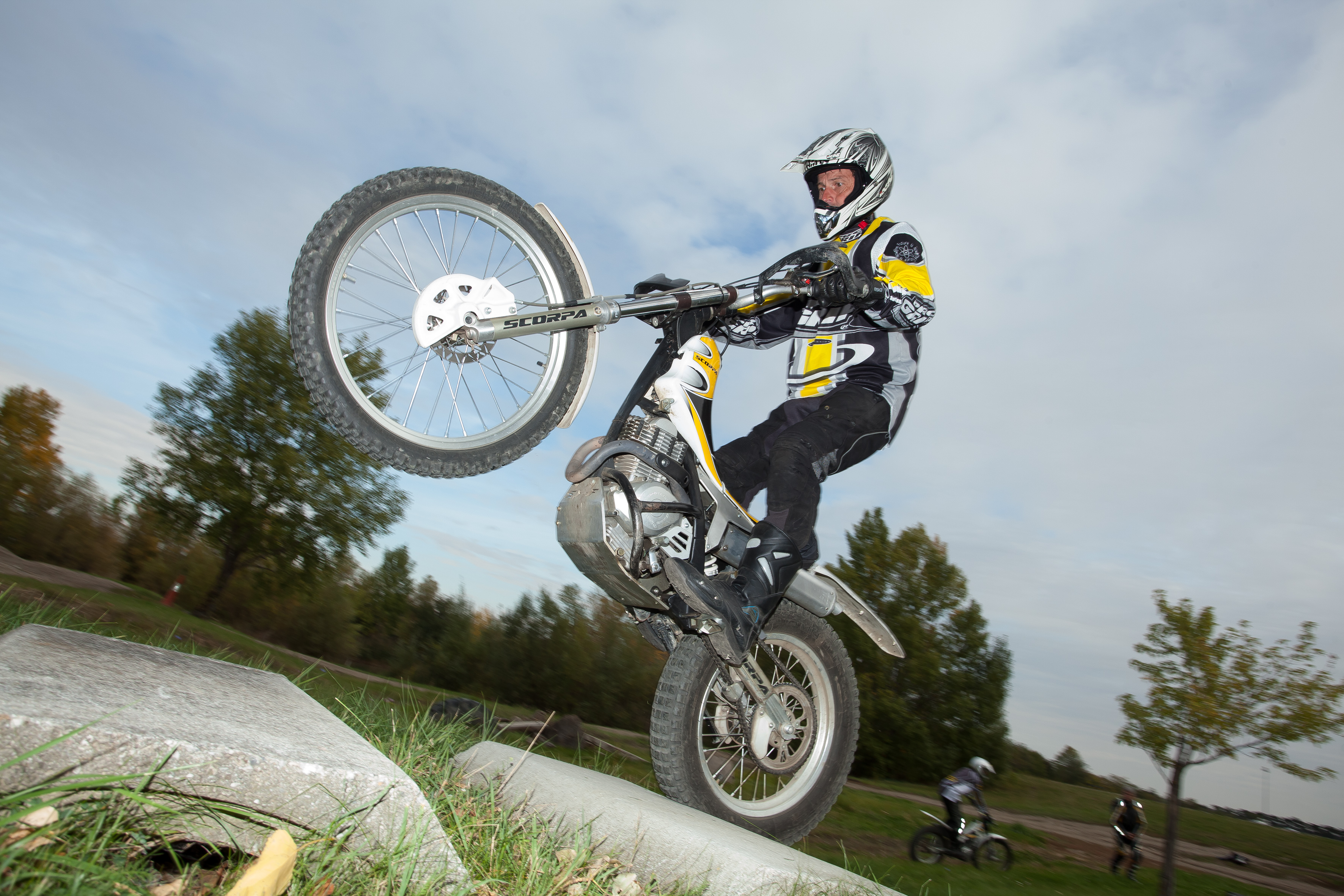 Eine Person mit Motorradhelm und Motorradkleidung fährt auf einem Trial auf dem Hinterreifen. Im Hintergrund sind blauer Himmel und Bäume.