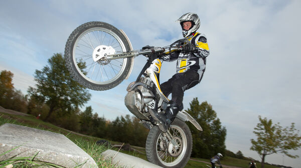 Eine Person mit Motorradhelm und Motorradkleidung fährt auf einem Trial auf dem Hinterreifen. Im Hintergrund sind blauer Himmel und Bäume.