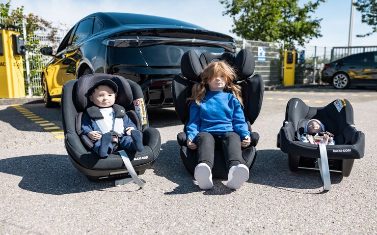 Vor einem Auto stehen drei Kindersitz mit Puppen drinnen.
