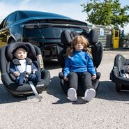 Vor einem Auto stehen drei Kindersitz mit Puppen drinnen.