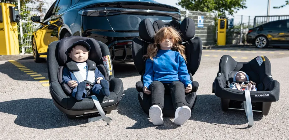 Vor einem Auto stehen drei Kindersitz mit Puppen drinnen.