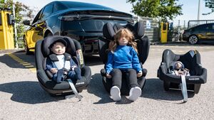 Vor einem Auto stehen drei Kindersitz mit Puppen drinnen.