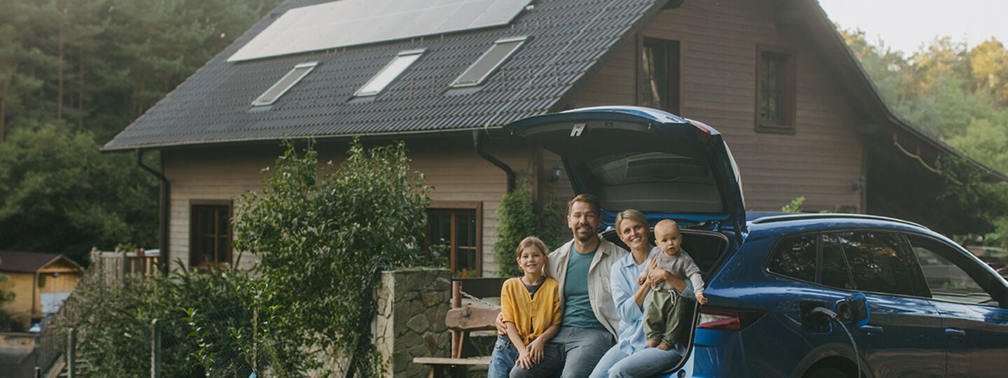 Vor einem Einfamilienhaus parkt ein Auto, im geöffneten Kofferraum sitzt eine Familie mit zwei Kindern. 