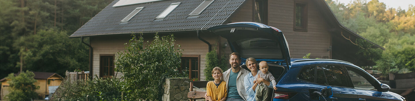 Vor einem Einfamilienhaus parkt ein Auto, im geöffneten Kofferraum sitzt eine Familie mit zwei Kindern. 