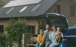 Vor einem Einfamilienhaus parkt ein Auto, im geöffneten Kofferraum sitzt eine Familie mit zwei Kindern.