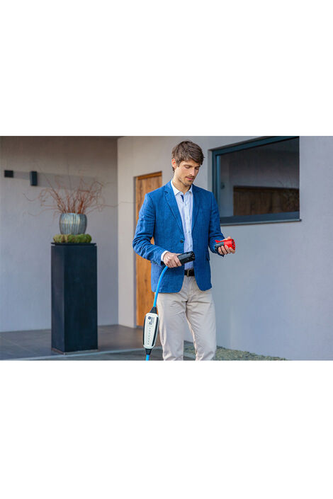 Ein Mann mit blauem Jacket steht vor einem Haus und hält ein Ladekabel für E-Autos in der Hand.