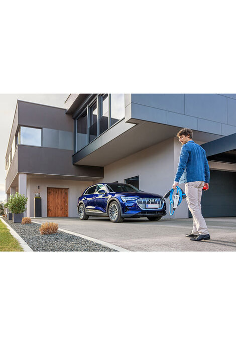 Ein modernes Wohnhaus, davor steht ein großes blaues Auto, ein Mann geht auf das Auto zu und hält ein Ladekabel in der linken Hand.