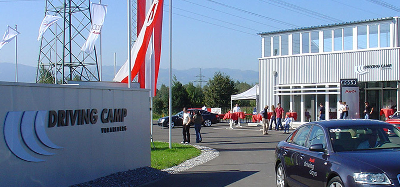Links ist eine Wand mit der Aufschrift „Driving Camp Vorarlberg“, rechts fährt ein schwarzes Auto. Im Hintergrund sind Personen, Fahnen, Strommasten, Autos und ein graues Gebäude.