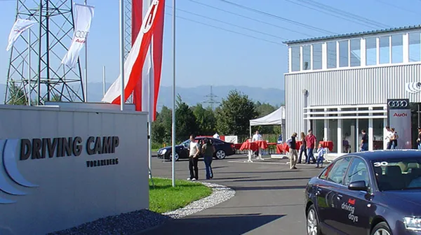 Links ist eine Wand mit der Aufschrift „Driving Camp Vorarlberg“, rechts fährt ein schwarzes Auto. Im Hintergrund sind Personen, Fahnen, Strommasten, Autos und ein graues Gebäude.