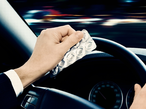 Das Bild zeigt die Hände von einem Autofahrer im Fahrzeug. Er sitzt am Steuer und hält mit einer Hand das Lenkrad, während er in der linken Hand ein Tablettenblister hält. 